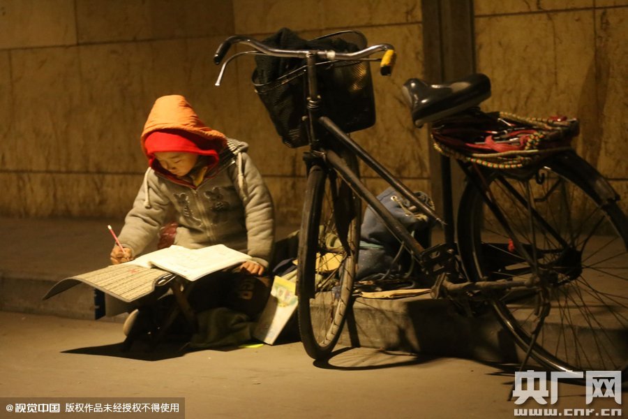鄭州中原路京廣鐵路大橋下涵洞的馬路牙子邊，一位戴著紅色辣椒帽的小男孩趴在一個(gè)小馬扎上，上面鋪著一張車(chē)墊，在微弱的燈光下寫(xiě)作業(yè)