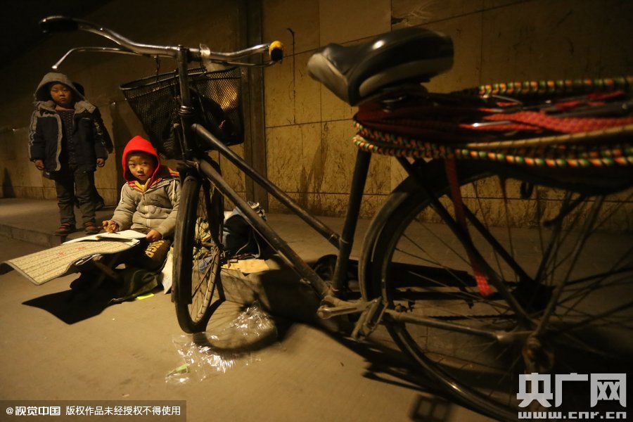 2015年11月8日晚，鄭州，7歲男孩涵洞橋下陪父母練攤，路邊燈下坐小馬扎上寫(xiě)作業(yè)。